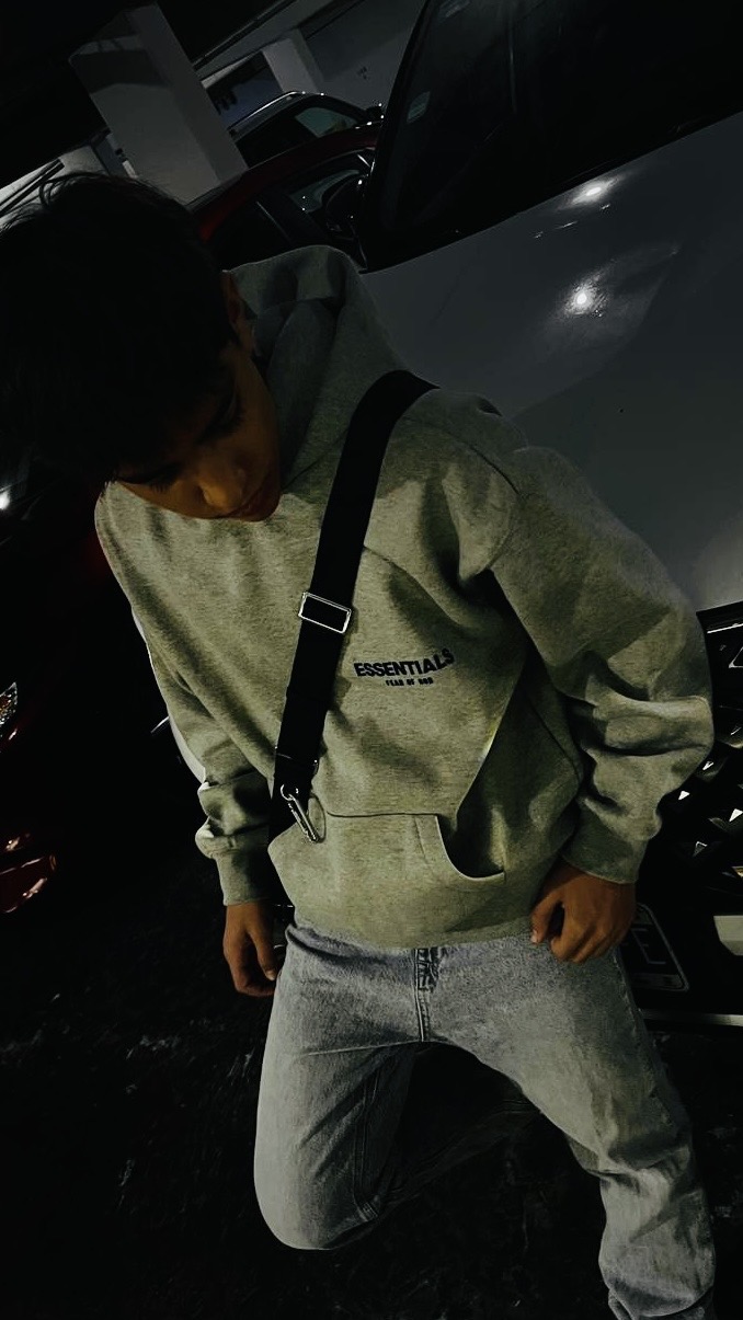 Persona usando hoodie Essentials gris con jeans claros en un estacionamiento.